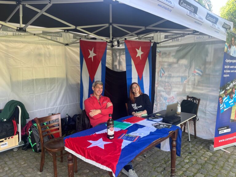 Zwei Personen sitzen an einem Tisch mit einer kubanischen Flagge. Hinter ihnen sind Zelte mit Werbebannern aufgestellt.