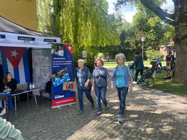 Drei ältere Frauen gehen nebeneinander auf einem Gehweg bei einem Charity-Event. Links im Bild steht ein Stand mit einer Kubaflagge und einem Banner, das für Spenden für Sportprojekte in Kuba wirbt.