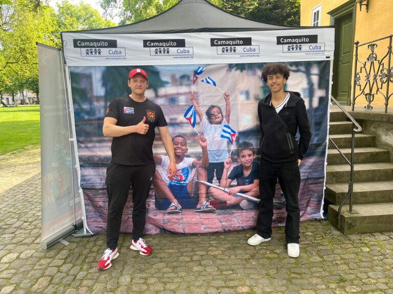 Zwei junge Männer stehen vor einem Pavillon mit einem Banner, auf dem Kinder mit kubanischen Flaggen abgebildet sind.