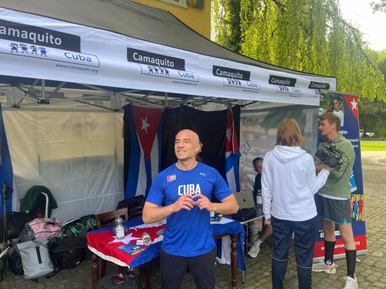 Ein Mann im blauem Sportshirt mit der Aufschrift 'Cuba' lächelt und posiert vor einem Informationsstand mit kuba-bezogener Dekoration. Hinter ihm stehen mehrere Personen in sportlicher Kleidung.