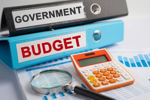 Eine Lupe und ein Taschenrechner liegen auf einem Tisch, dahinter zwei Aktenordner mit der Aufschrift "Government" und "Budget"