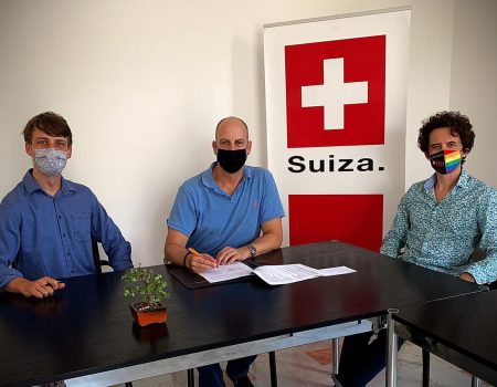 Drei Personen sitzen an einem Tisch, im Hintergrund das Schweizer Wappen